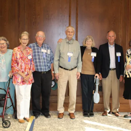 Group of seven smiling elderly people standing indoors against wood panel wall, some using walking aids and nametags.