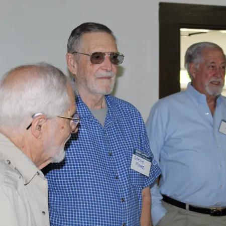 Three elderly men wearing name tags, casually standing and conversing in a light, indoor room with stone walls.