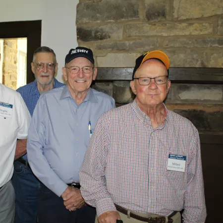 Four elderly men standing indoors near a fireplace, wearing glasses and name tags, posing for a group photo.