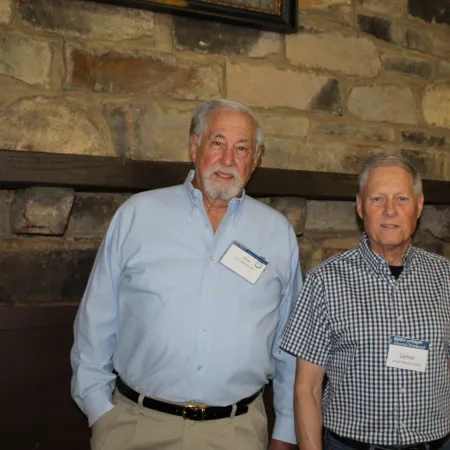 Two older men with name tags standing indoors against a stone wall and window background.
