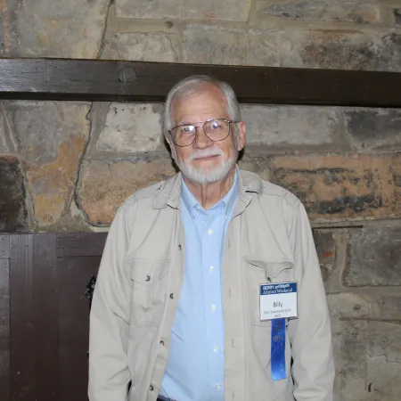 Elderly man wearing glasses and name tag smiling in front of stone wall with wooden beam.