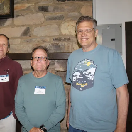 Four middle-aged men standing indoors against a stone wall, two wearing glasses and name tags, smiling at the camera.