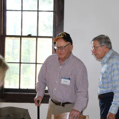 Two elderly men in checkered shirts talking indoors near a window, one with a cap and cane, the other wearing glasses.