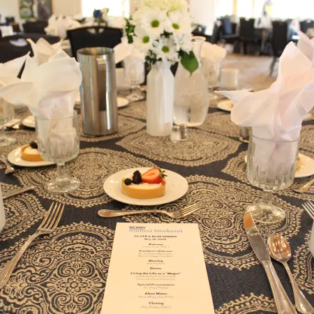 Elegant banquet table set with patterned tablecloth, white napkins, floral centerpiece, and dessert plates.