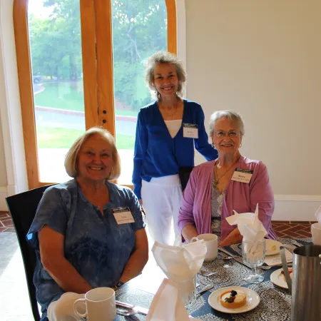 Four smiling elderly women gathered around a dining table set with flowers, plates, and beverages in a bright room.