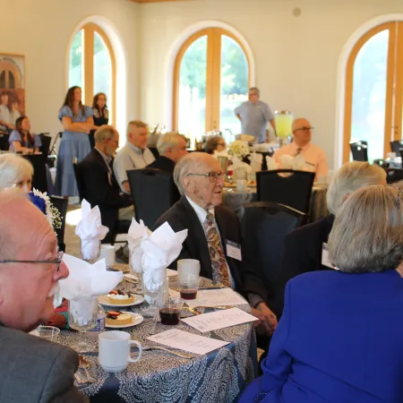 Group of elderly people and guests seated at tables in a bright room with arched windows during an event.