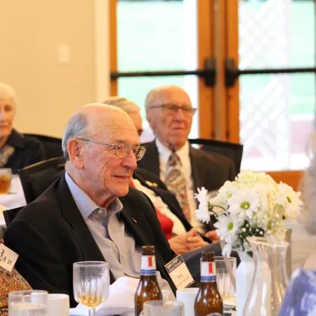 Elderly people in a formal gathering sitting at a decorated table with drinks and flowers indoors.