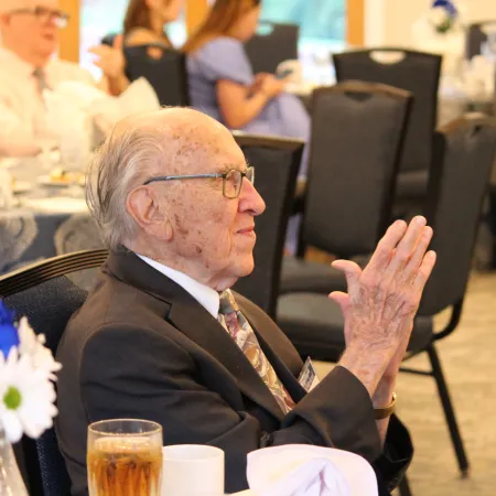 Elderly man in suit clapping hands at formal indoor event with other attendees seated in background.