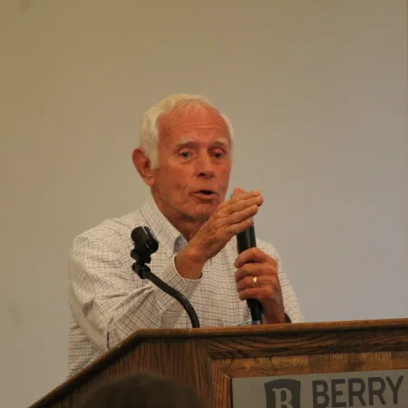 Elderly man speaking into a microphone behind a wooden podium labeled Berry in an indoor setting.
