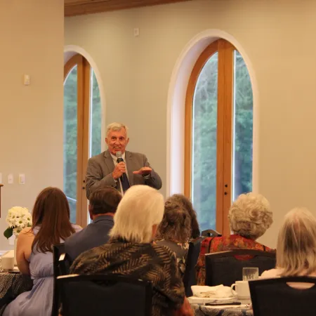 Man speaking with microphone to seated audience in a bright room with arched windows and dining setup