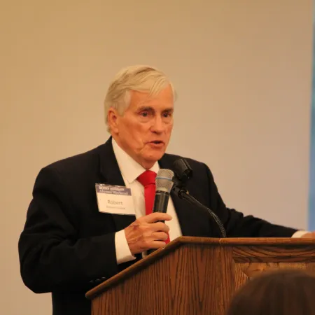 Older man with gray hair in suit speaking into microphone at wooden podium during indoor event