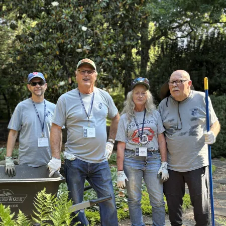Four volunteers wearing gloves and work shirts stand outdoors with gardening tools and wheelbarrow by green plants.
