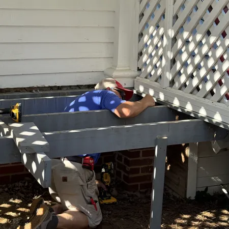 Man in blue shirt and cap kneeling while repairing a wooden deck attached to a house under sunlight.