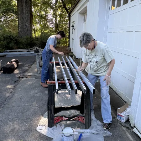 Two people painting long wooden slats outside near garage with paint cans and brushes on plastic sheet.