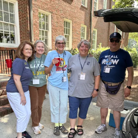 Group of five adults smiling outside a brick building during a casual gathering on a sunny day.