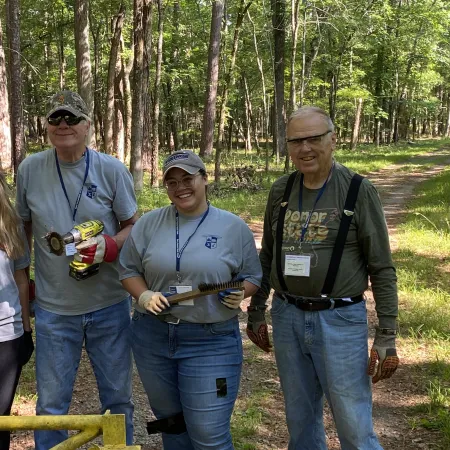 Four volunteers in a forest trail clearing with tools, smiling and wearing gloves and casual work clothes.
