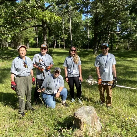 Group of five volunteers in gray shirts standing in a grassy, wooded area with landscaping tools on a sunny day