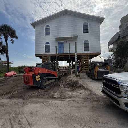 Two-story house elevated on stilts with construction equipment and a pickup truck nearby under a cloudy sky.