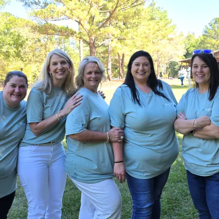 Five women in matching light blue t-shirts pose outdoors on a sunny day with trees and grass in the background.