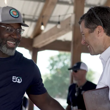 Two men smiling and shaking hands at an outdoor event under a wooden pavilion.