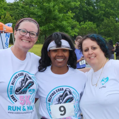 Three smiling women wearing matching 5K run event shirts posing outdoors by a finish line banner.