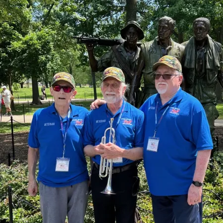 Three veterans in blue shirts and hats pose in front of the Vietnam Veterans Memorial statue in a sunny park.