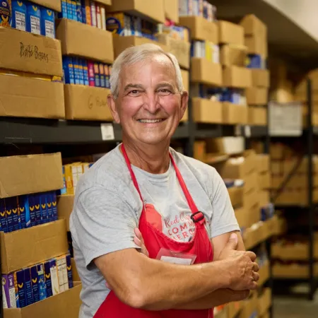 Smiling older man in red apron standing with arms crossed in a pantry filled with boxed food items.