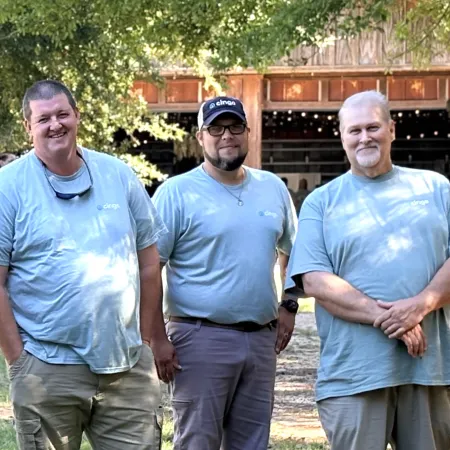 Three men in matching light blue shirts stand outdoors in front of a wooden barn with balloons nearby on a sunny day