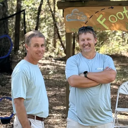 Two men in light blue shirts and light pants smiling outdoors near a wooden sign with the word Foot