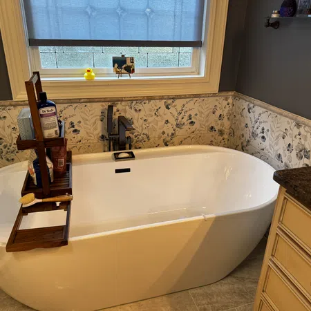 Modern white freestanding bathtub with wooden caddy, floral wallpaper, blue window shade, and granite countertop.