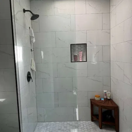 Modern walk-in shower with white hex tile floor, large marble wall tiles, glass door, and wooden stool with toiletries.