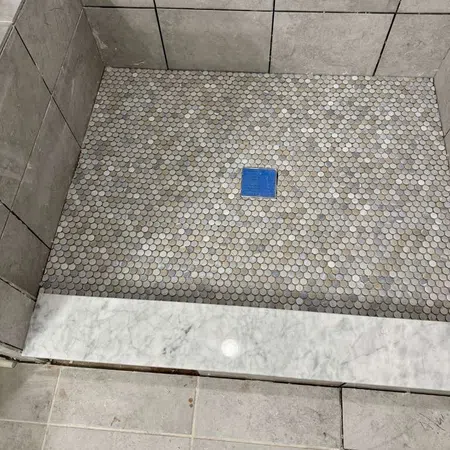 Gray tiled shower floor with small circular mosaic tiles and blue drain cover, surrounded by large gray wall tiles.