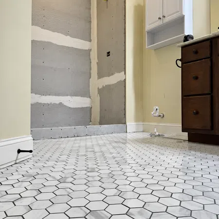 Bathroom under renovation with white hexagonal marble floor tiles, beige walls, and wooden cabinet.