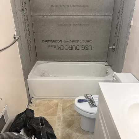 Bathroom under renovation with cement board around bathtub, beige floor tiles, toilet, and white vanity cabinet.