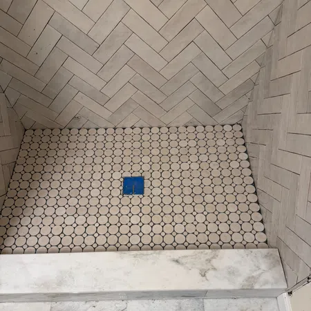 Shower floor with round white tiles and blue drain surrounded by beige herringbone patterned wall tiles.