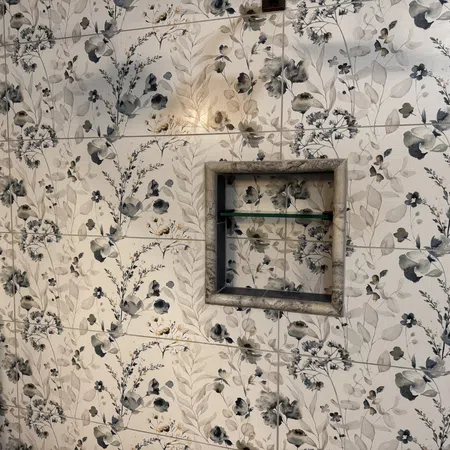 Tiled bathroom wall with floral pattern in shades of gray, beige, and blue featuring a recessed shelf with glass.