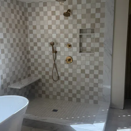 Modern shower with beige checkered tiles, gold fixtures, marble bench and a white freestanding bathtub.