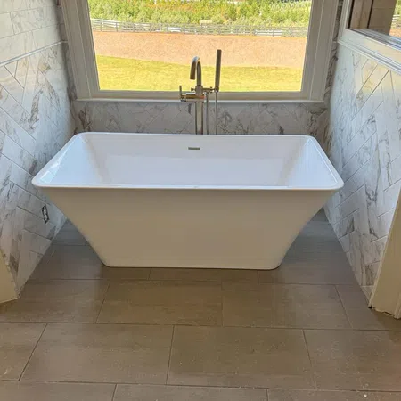 White freestanding bathtub with floor-mounted faucet in a tiled bathroom with a large window overlooking greenery.
