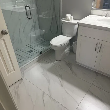 Modern bathroom with marble floor tiles, glass shower, white toilet, and white vanity under natural light.