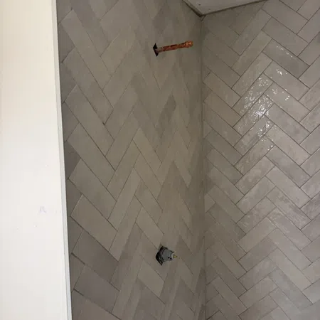 Gray herringbone tile wall in a corner with exposed plumbing pipes for a shower installation.