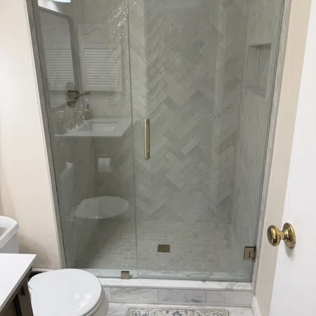 Modern bathroom with glass shower door, herringbone tile pattern, white toilet, and decorative rug on marble floor.