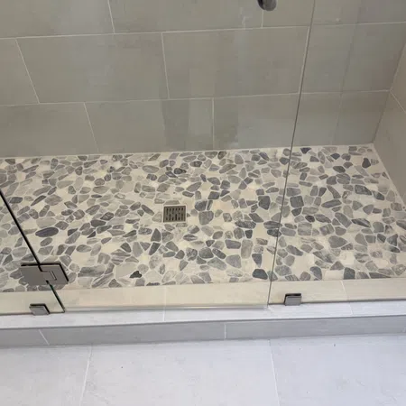 Modern shower area with pebble stone floor tiles and glass door enclosure with metal hinges and drain.