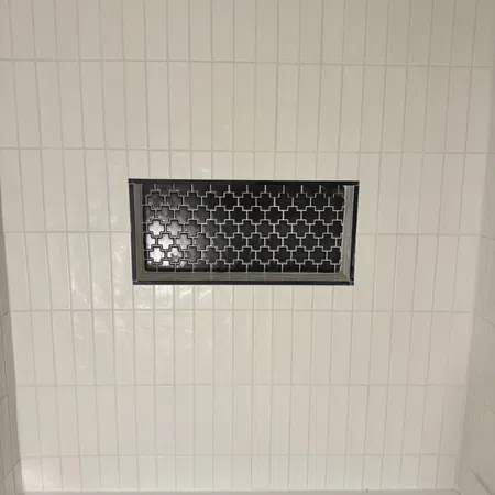 White tiled bathroom wall with a decorative black vent grate above a white bathtub