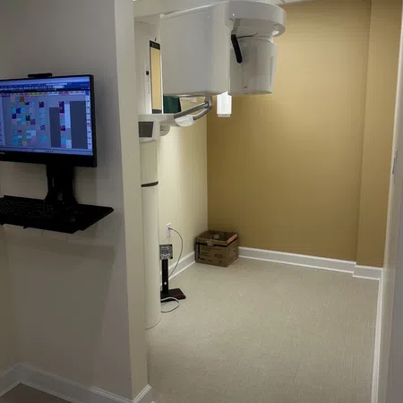 Modern dental X-ray machine in a clean beige and white clinic room with a wall-mounted computer screen.