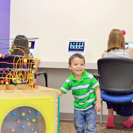 Young boy in green striped shirt playing near colorful bead maze toy in a bright pediatric dentistry waiting room.