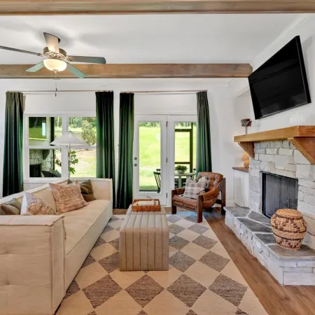 Bright living room with beige sofa, stone fireplace, patterned rug, green curtains, and wooden ceiling beams