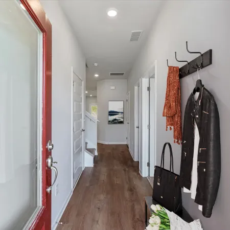 Modern entryway with wooden floor, red door, coat rack with jacket and scarf, and decorative flowers on bench.