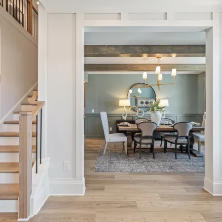 View of modern dining room with wooden beams, gray walls, chandelier, and light wood staircase and flooring.