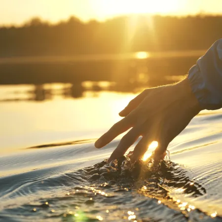 A hand gently touches the water's surface during sunset, creating ripples in a serene landscape.