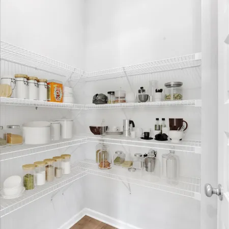Organized pantry with white wire shelves holding jars, containers, dishes, and kitchen essentials on light wood flooring.